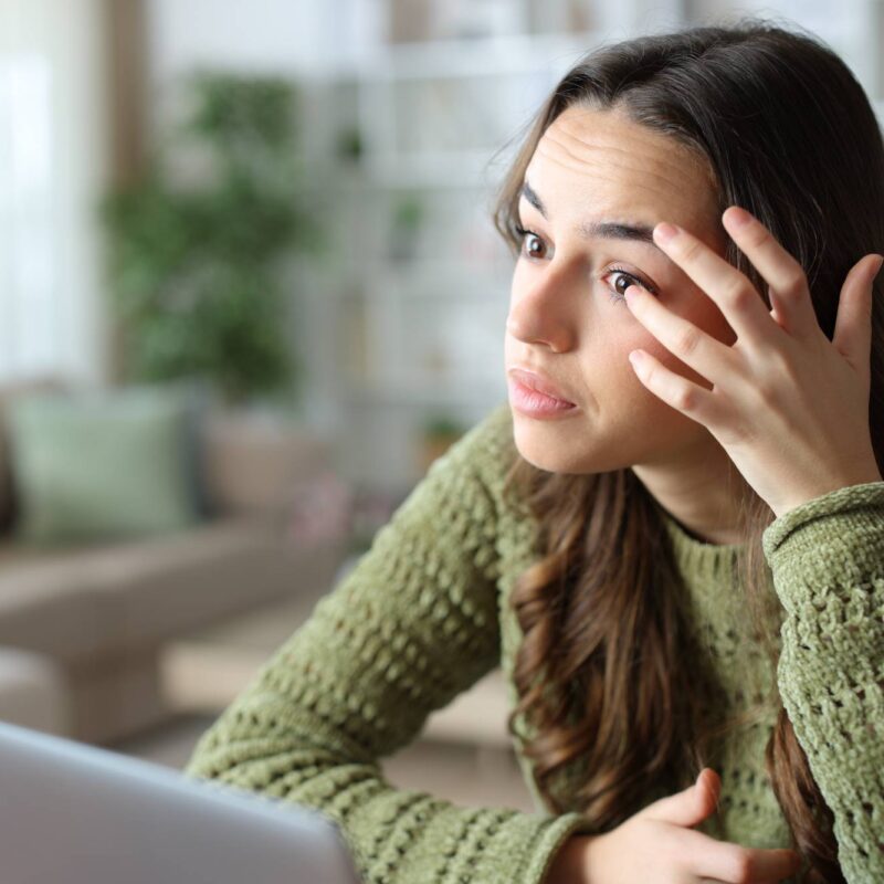 Young woman touching her eye in discomfort at home, experiencing dry eye symptoms from contact lens wear